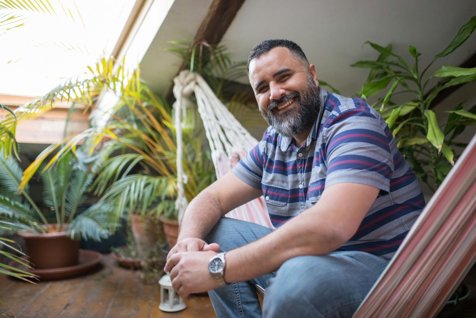 Man in a striped shirt relaxing in a hammock indoors with lush plants around.